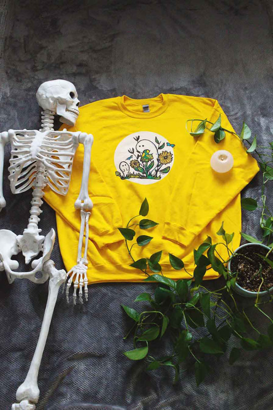 Photo of gold sweatshirt with ghosts and florals on it from top view pictured with a skeleton and flowers
